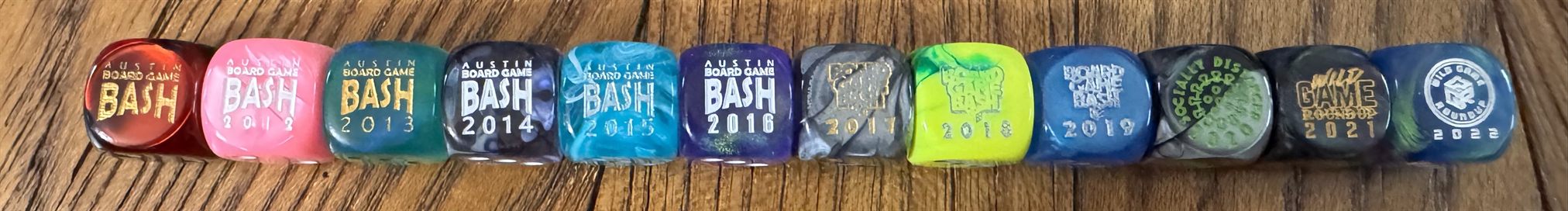 Twelve years of Board Game Bash custom dice lined up in order, 2011 through 2022